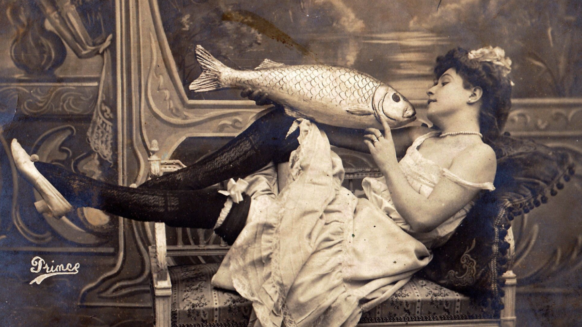 A vintage black & white photo shows a woman lounging on a bench holding a large fake fish up to her face